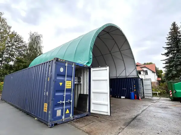Tymczasowa hala z kontenerów morskich, z zielonym, łukowatym dachem, chroni sprzęt przed deszczem.