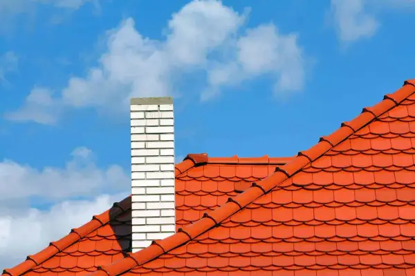 Pomarańczowy dach wielospadowy z ceglanym kominem na tle błękitnego nieba z chmurami.