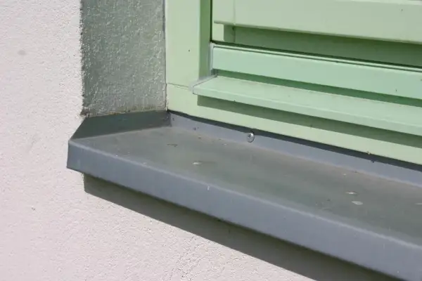 Szary parapet zewnętrzny zamontowany przy oknie z zielonymi okiennicami. Idealny moment, kiedy montować parapety zewnętrzne, to po ociepleniu i otynkowaniu elewacji.